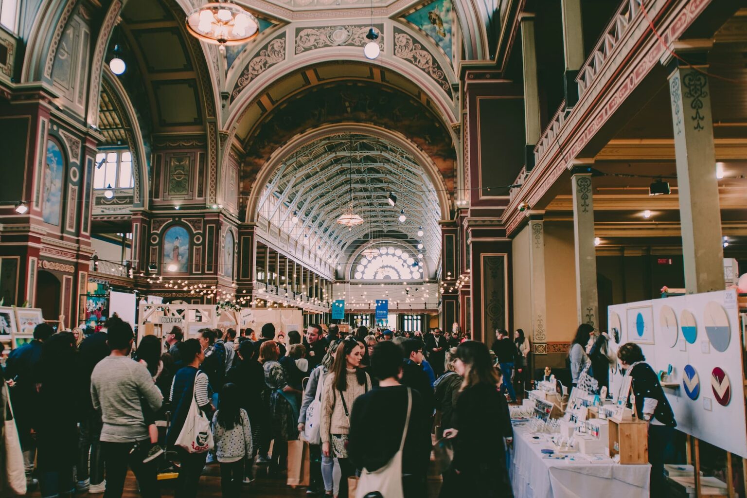 Grand event hall filled with attendees interacting with exhibitors and participating in activities. The hall features ornate architectural details and a lively atmosphere.